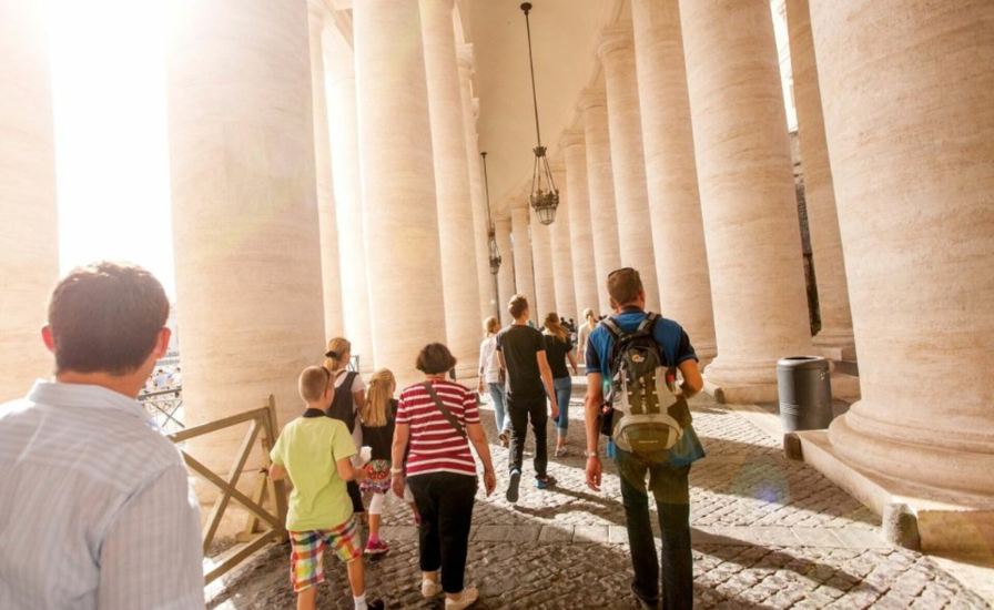 A group of people walking down a walkway with pillars, as they climb on St Peter Basilica
