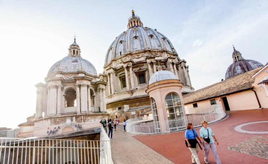 Tourists walking along Piazza del Santuario in Rome, with the iconic St. Peter's Basilica dome in the background
