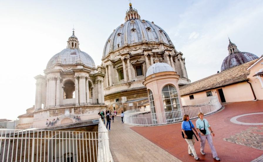 Tourists walking on Piazza del Santuario in Rome, with a view of St. Peter's Basilica in the background.
